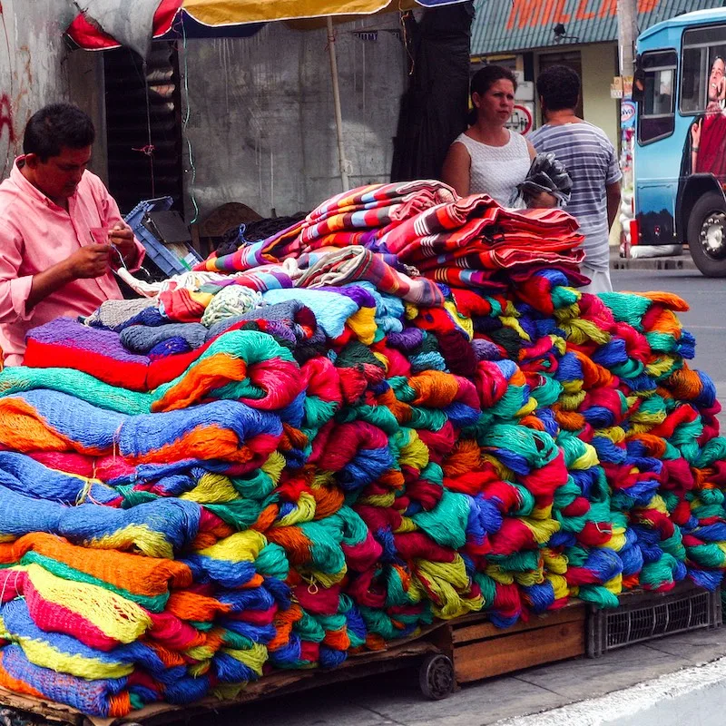 A sidewalk street vendor waits for a customer to sell one of his colorful hammocks to passersby on a busy street San Salvador.