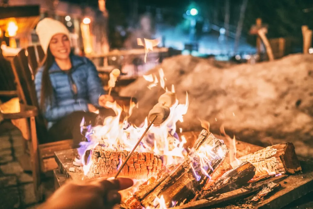 Winter holiday ski resort woman roasting marshmallows in BBQ firepit afterski fun leisure activity with friends. Couple grilling marshmallow stick in fire.